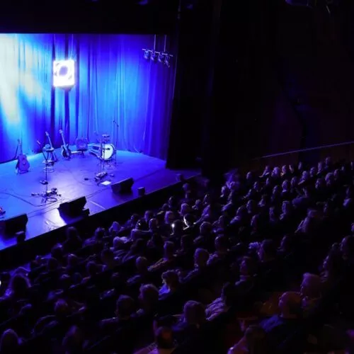 Aufnahme der leeren Bühne in blauem Licht mit vollbesetzten Sitzreihen.