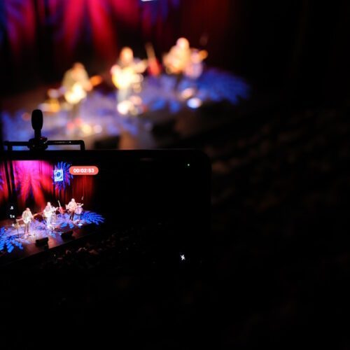 Aufnahme eines Handy-Displays, das die Band auf der Bühne zeigt mit der Bühne unscharf im Hintergrund.