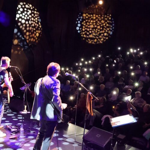 Die Band von hinten während des Auftritts im Scheinwerferlicht. Zahlreiche Handy-Taschenlampen im Zuschauerraum.